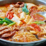 Budae jjigae being boiled in a large skillet with a spoon in the broth